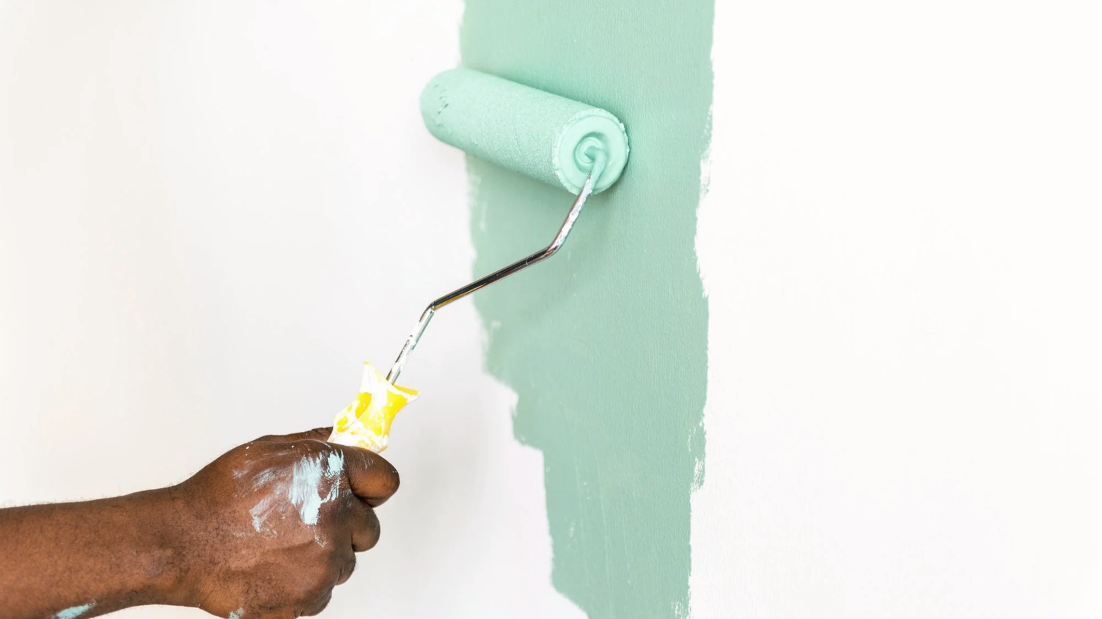 Hand with paint roller applying soft mint green paint on white wall for home renovation.