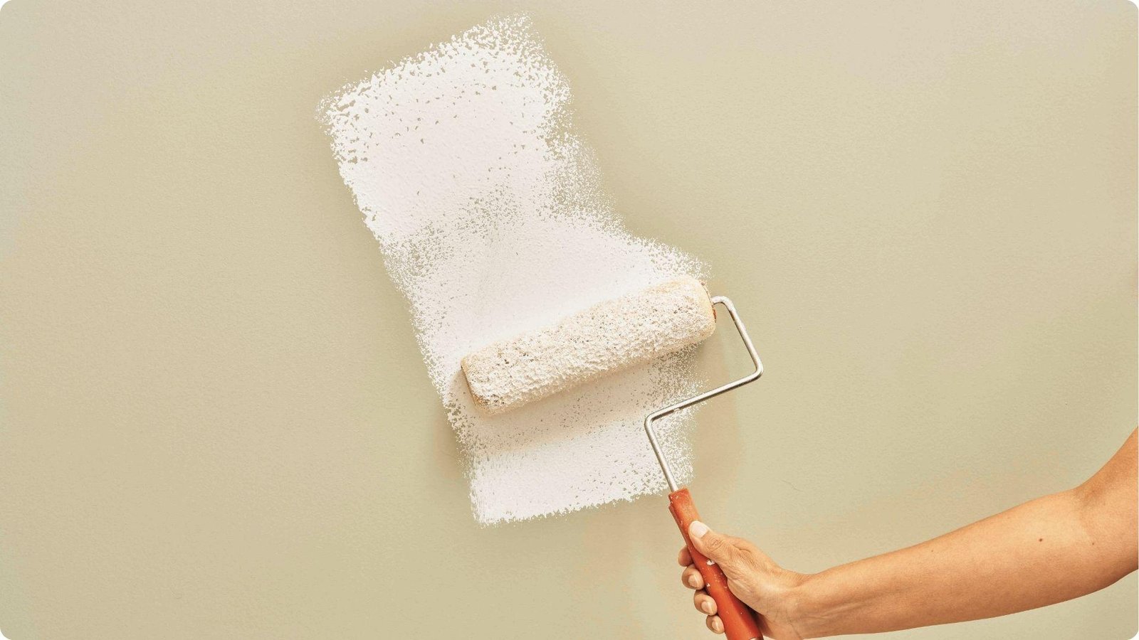 A hand rolls white paint onto a beige wall, showing the textured, partial coverage of a single pass.