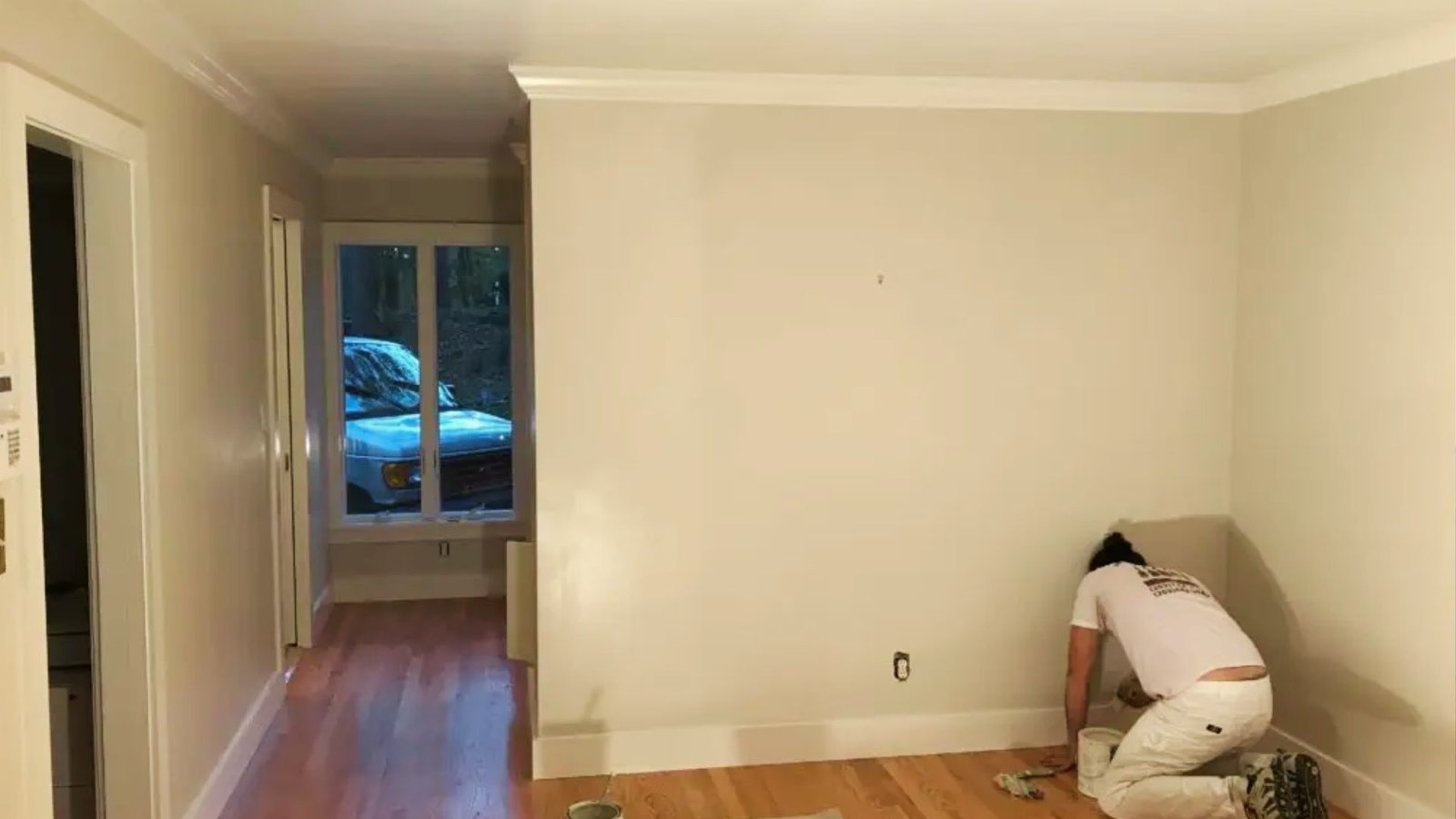 Professional painter applying white semi-gloss trim paint to baseboards in living room during home interior renovation