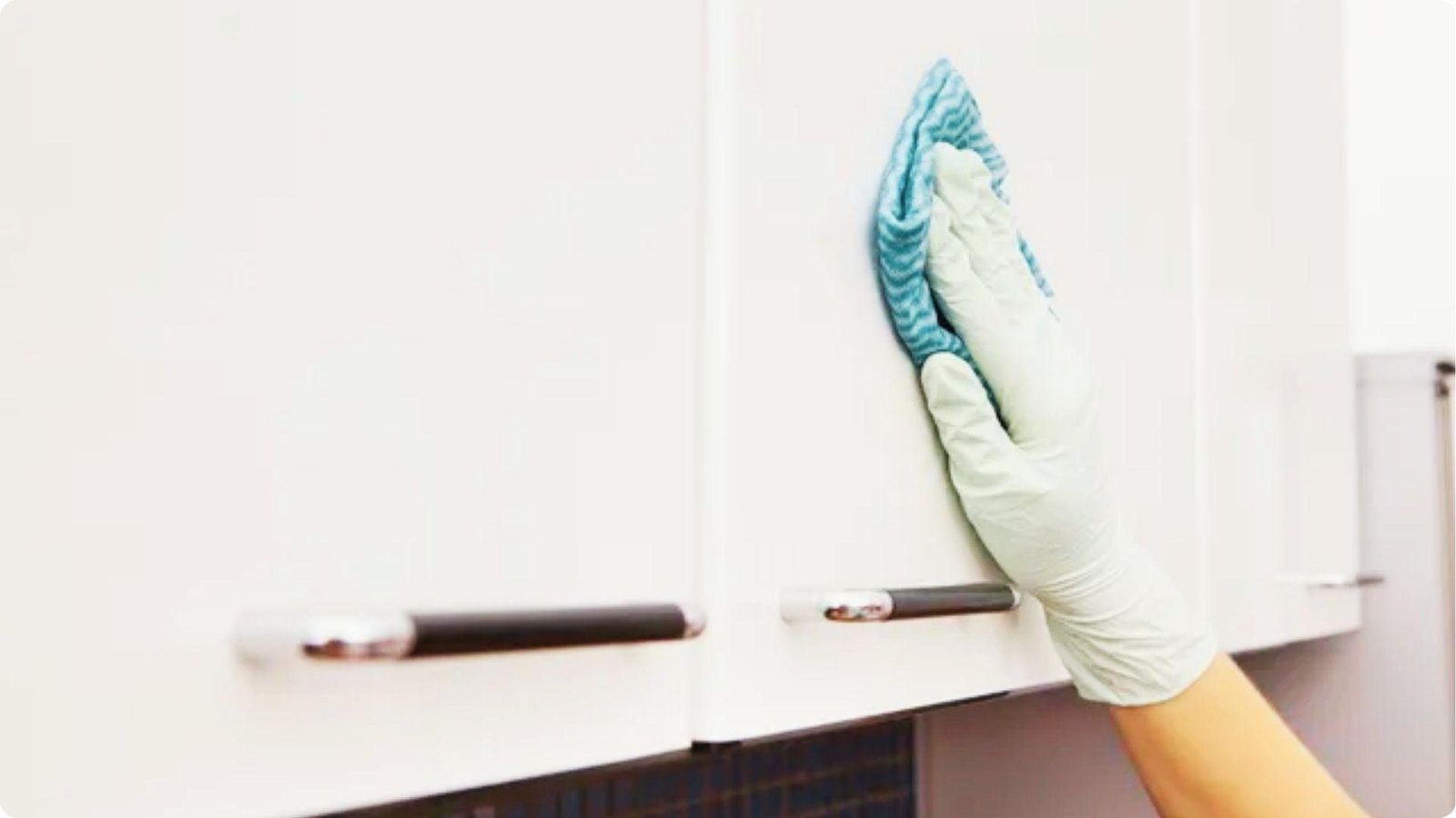 Close-up of a gloved hand using a blue striped cloth to clean the surface of a white painted cabinet door.