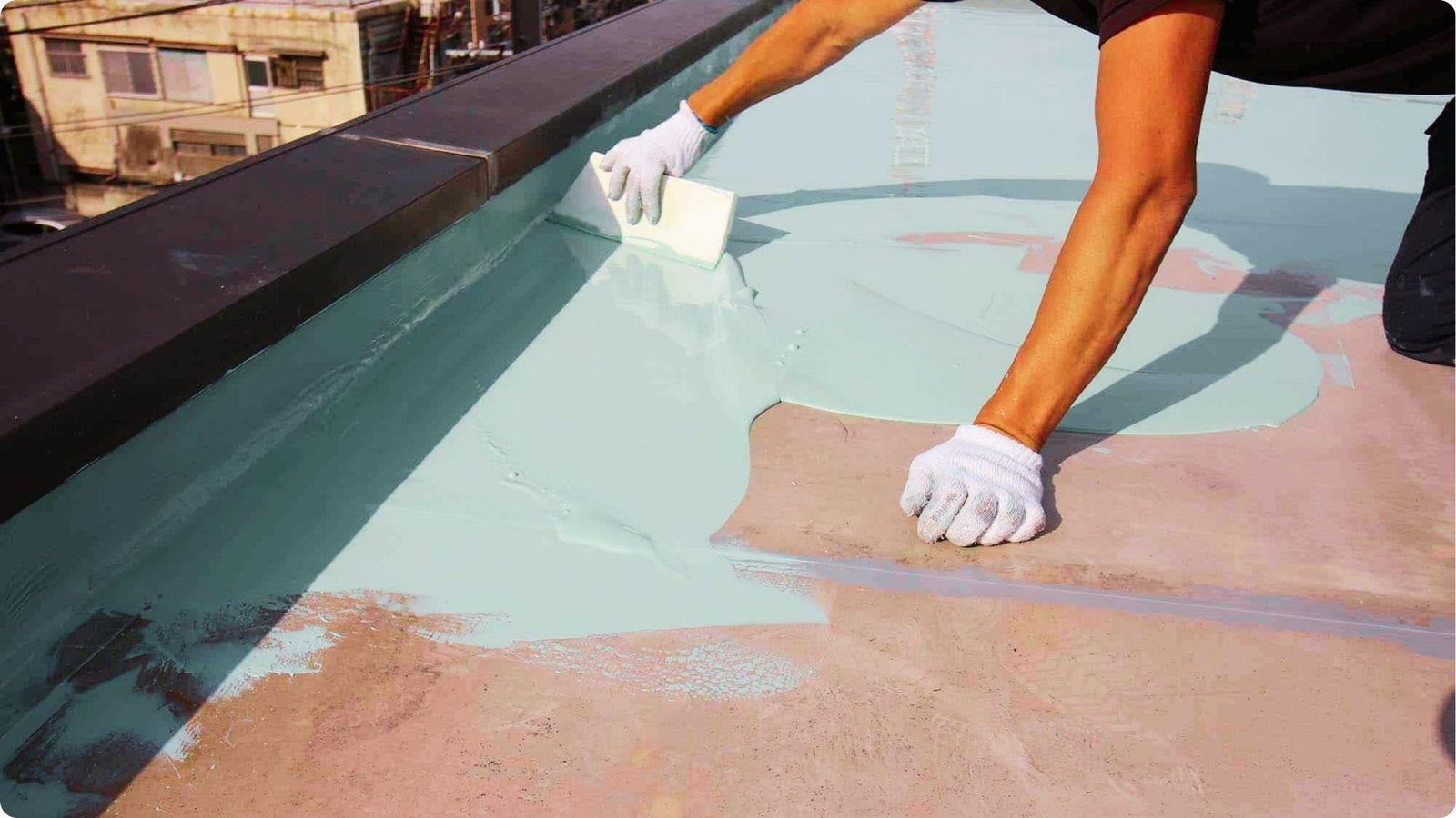 Worker wearing white gloves applies light blue roof coating to a flat TPO surface with a trowel near the parapet wall.
