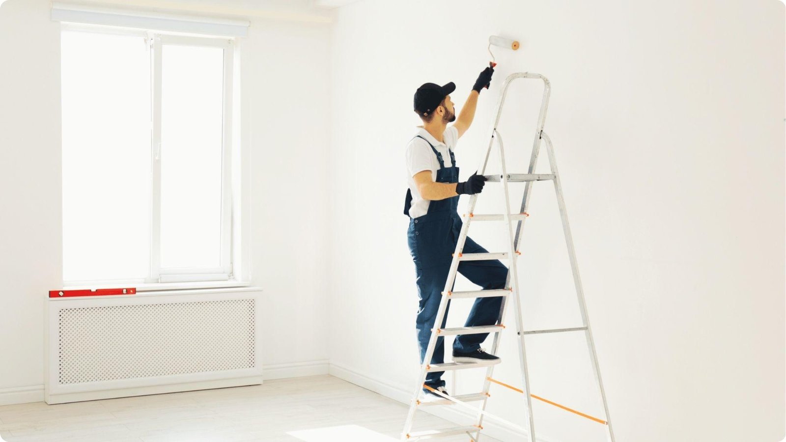 Painter in overalls on a ladder applying white paint near the ceiling of a bright room.