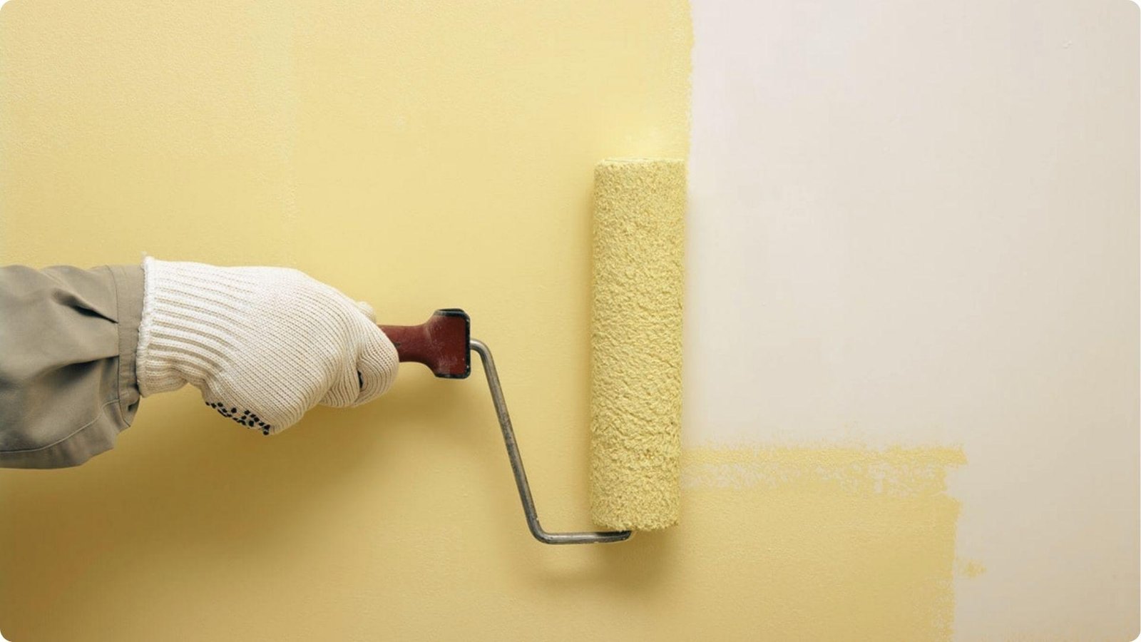 A gloved hand rolls yellow paint onto a white wall, creating a sharp line between the old and new color.