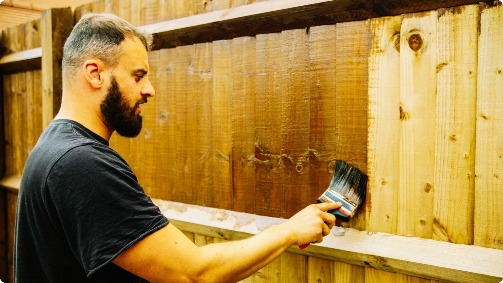A man with a beard uses a large brush to apply brown stain to a vertical wood fence.