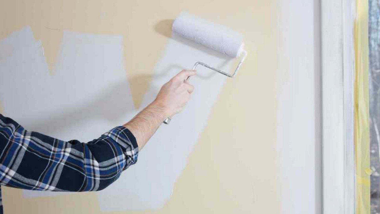 Person rolling white paint over beige wall, waiting for paint to dry between coats prevents peeling