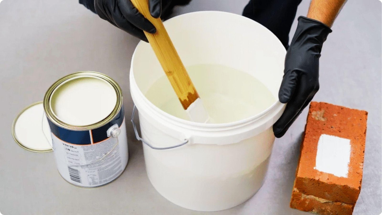 Gloved hands stir white paint and water in a bucket to create the whitewash mixture.