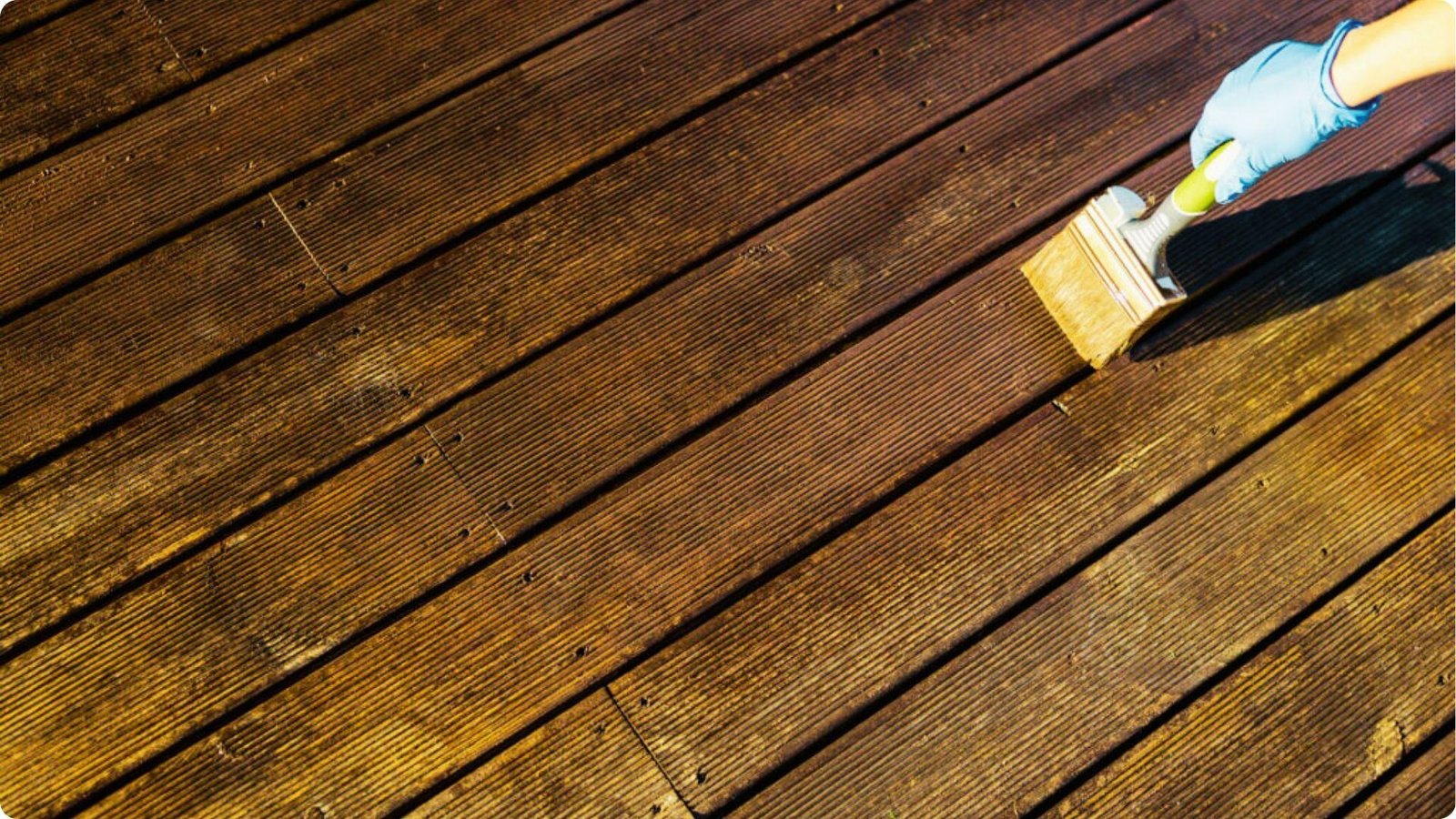 A gloved hand paints dark brown stain on grooved deck boards using a wide brush.