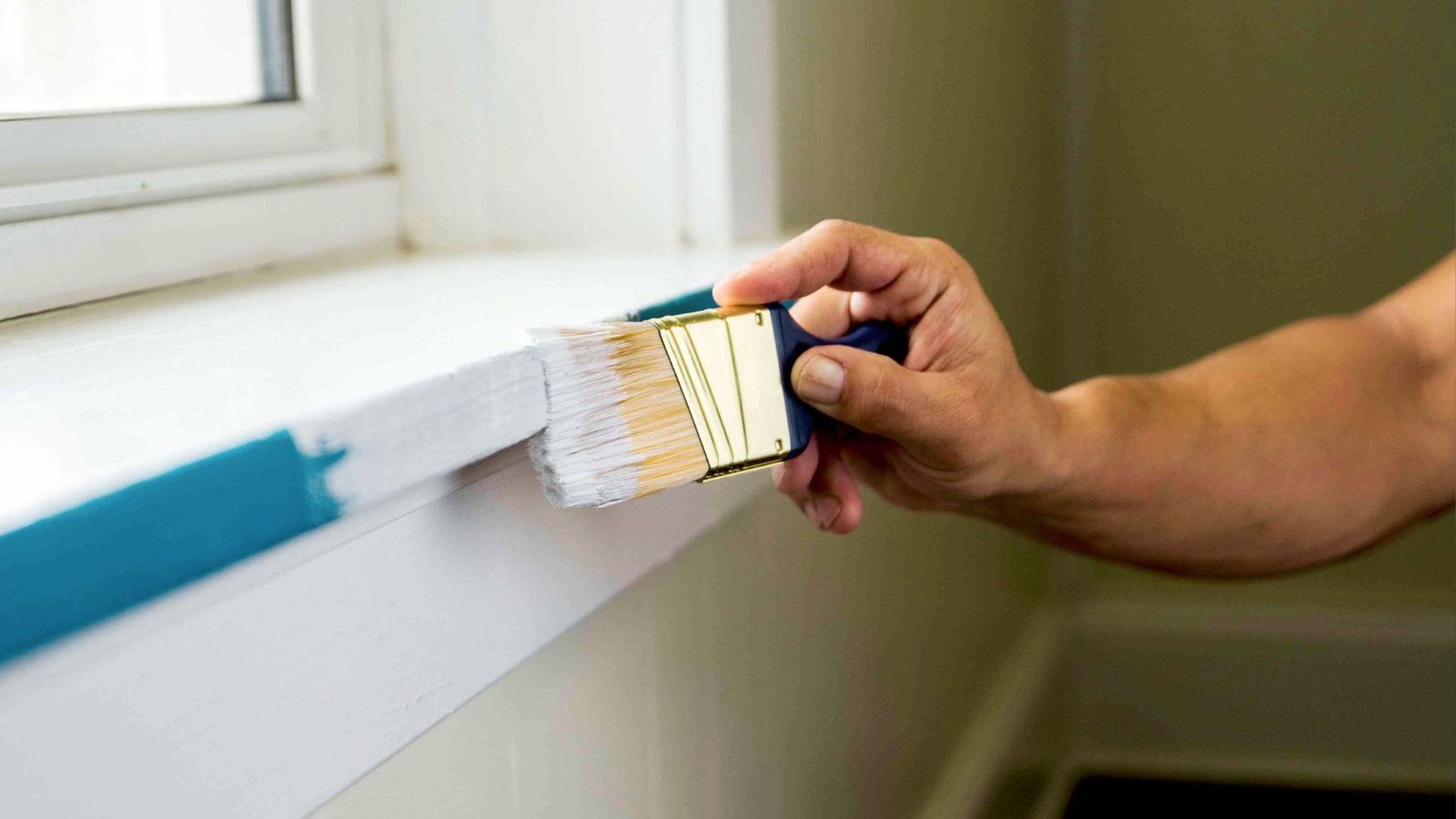 Hand painting white trim paint on window sill with blue painter's tape showing DIY home improvement and trim work