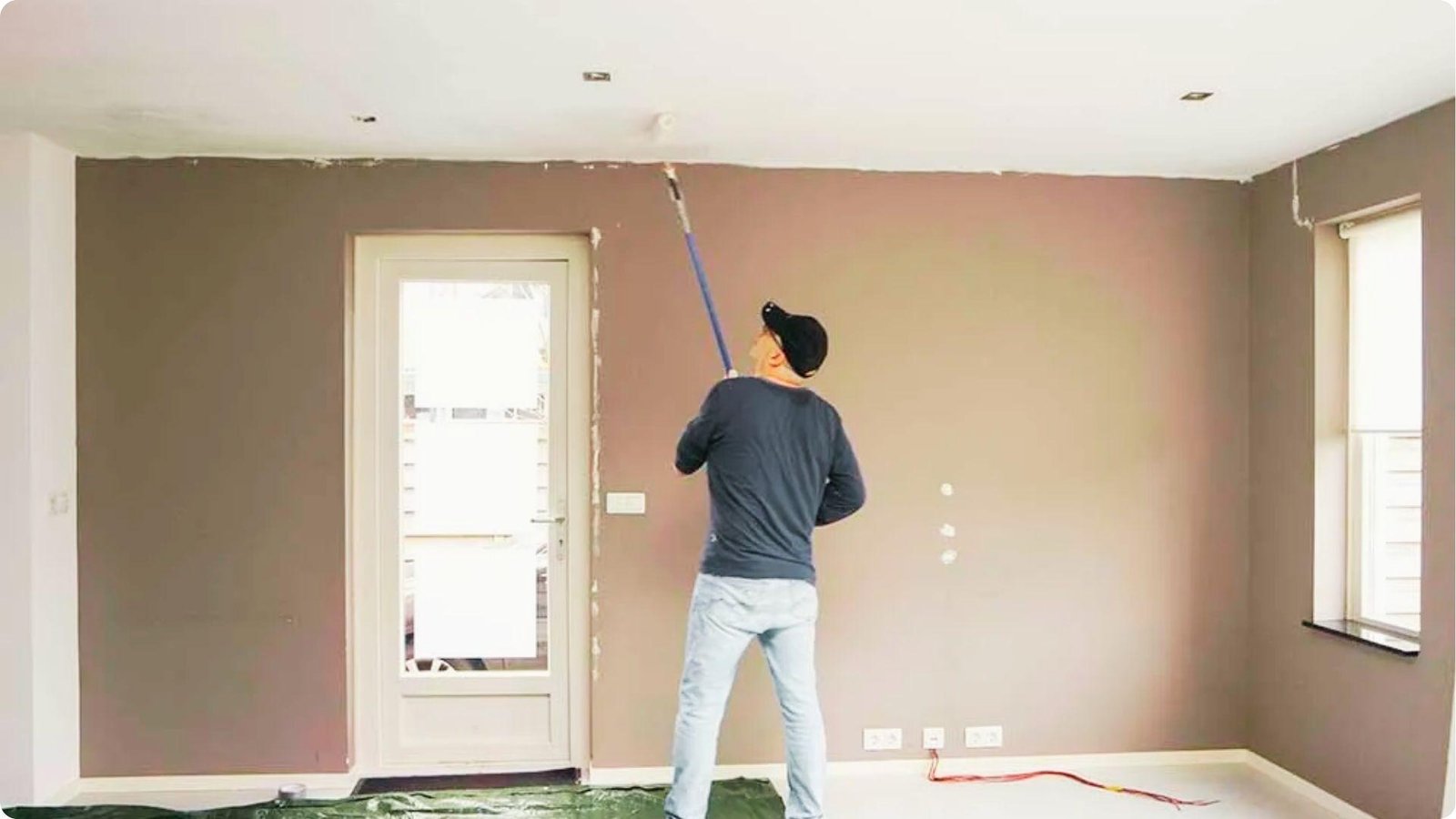 Man in a black shirt and cap uses an extension roller to paint a ceiling while brown walls surround him.
