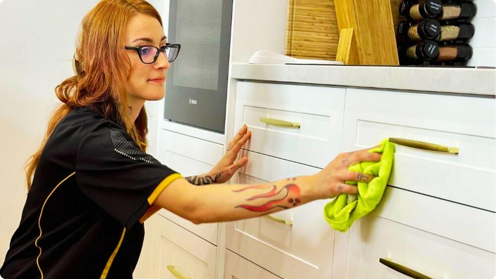 A woman with glasses and arm tattoos wipes white painted kitchen cabinets using a bright green microfiber cloth.