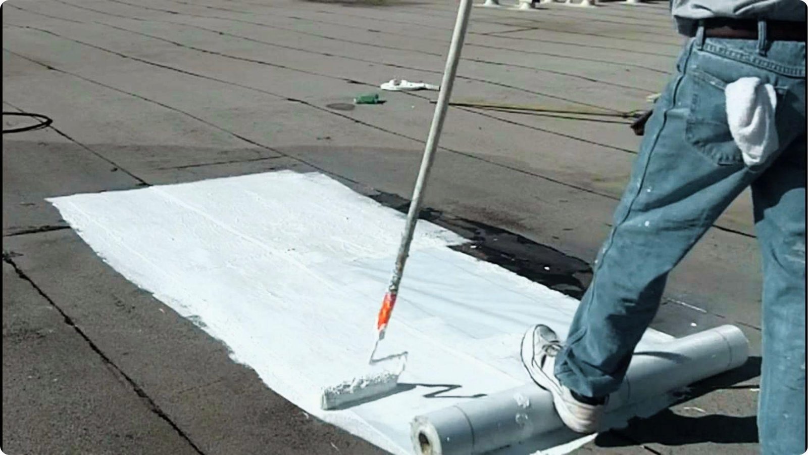 Worker rolls white reflective coating over a flat commercial roof next to a membrane roll.
