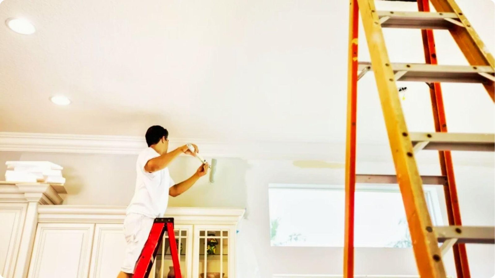Painter on a ladder carefully cutting in paint along the ceiling line in a bright room.
