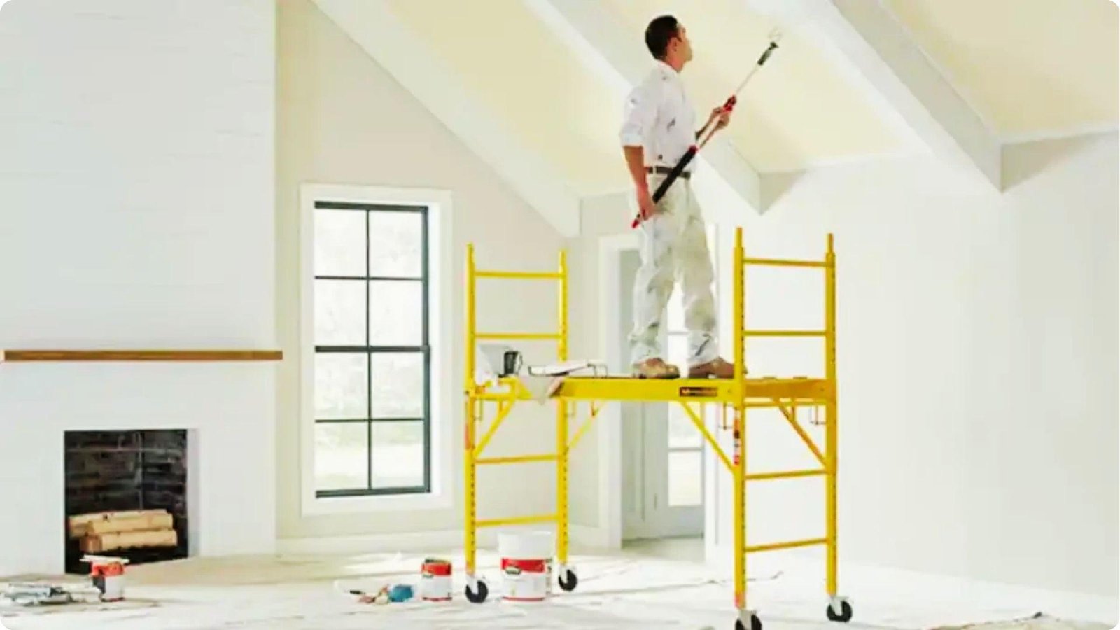 Painter on yellow scaffolding rolls paint on a high ceiling in a bright living room with a fireplace.
