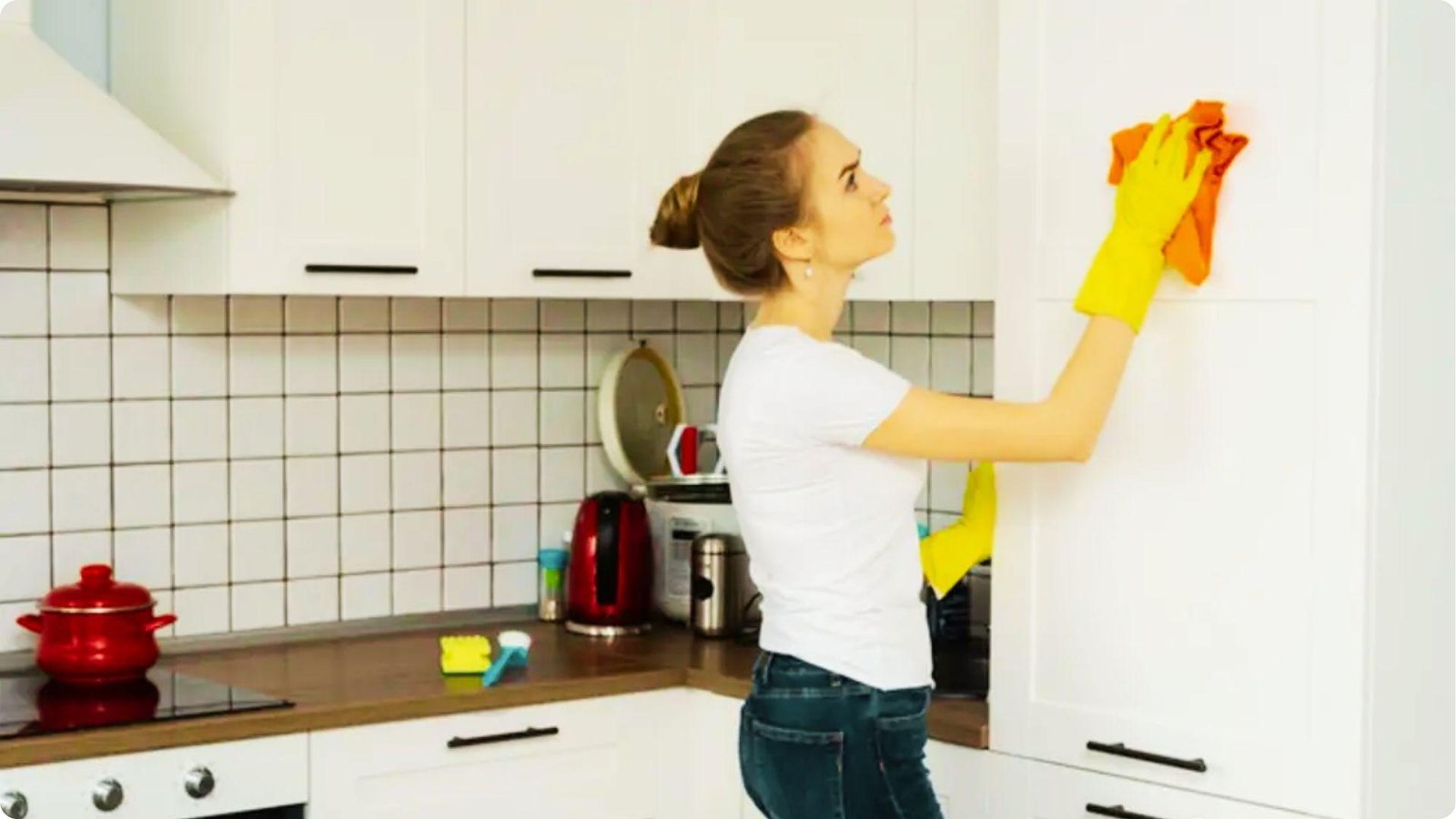 Woman in yellow gloves and white shirt cleans white upper kitchen cabinets with an orange cloth.