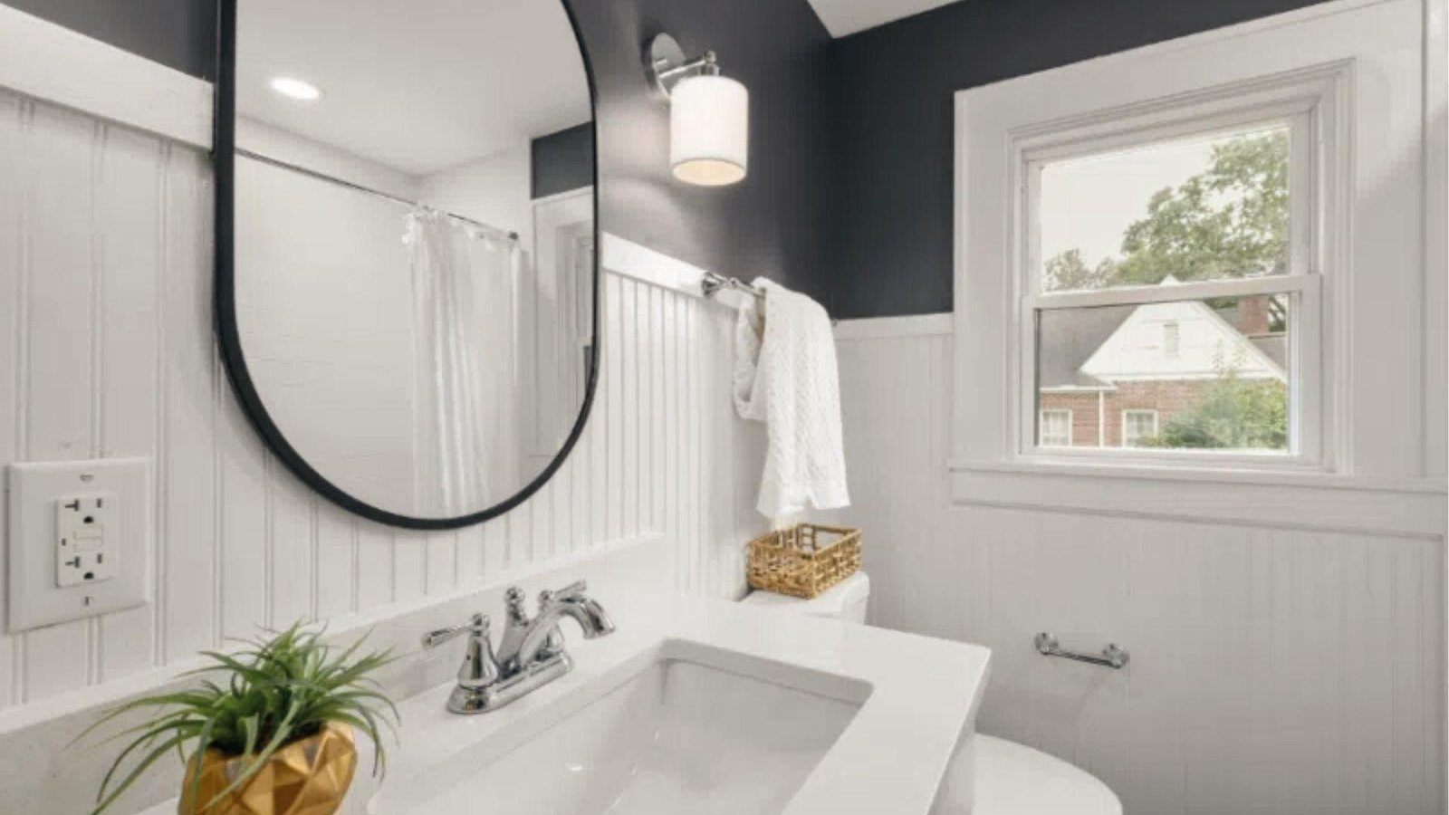 Bathroom with black upper wall and white wainscoting below oval mirror and gold planter.