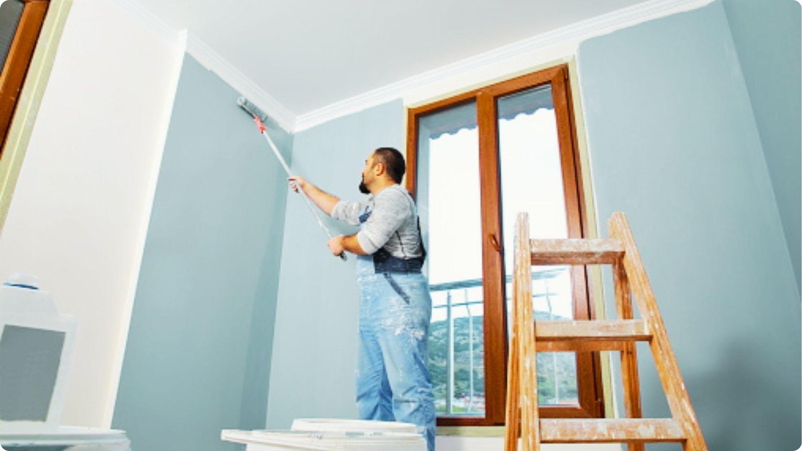 Contractor in overalls rolling light blue paint near crown molding with ladder setup.