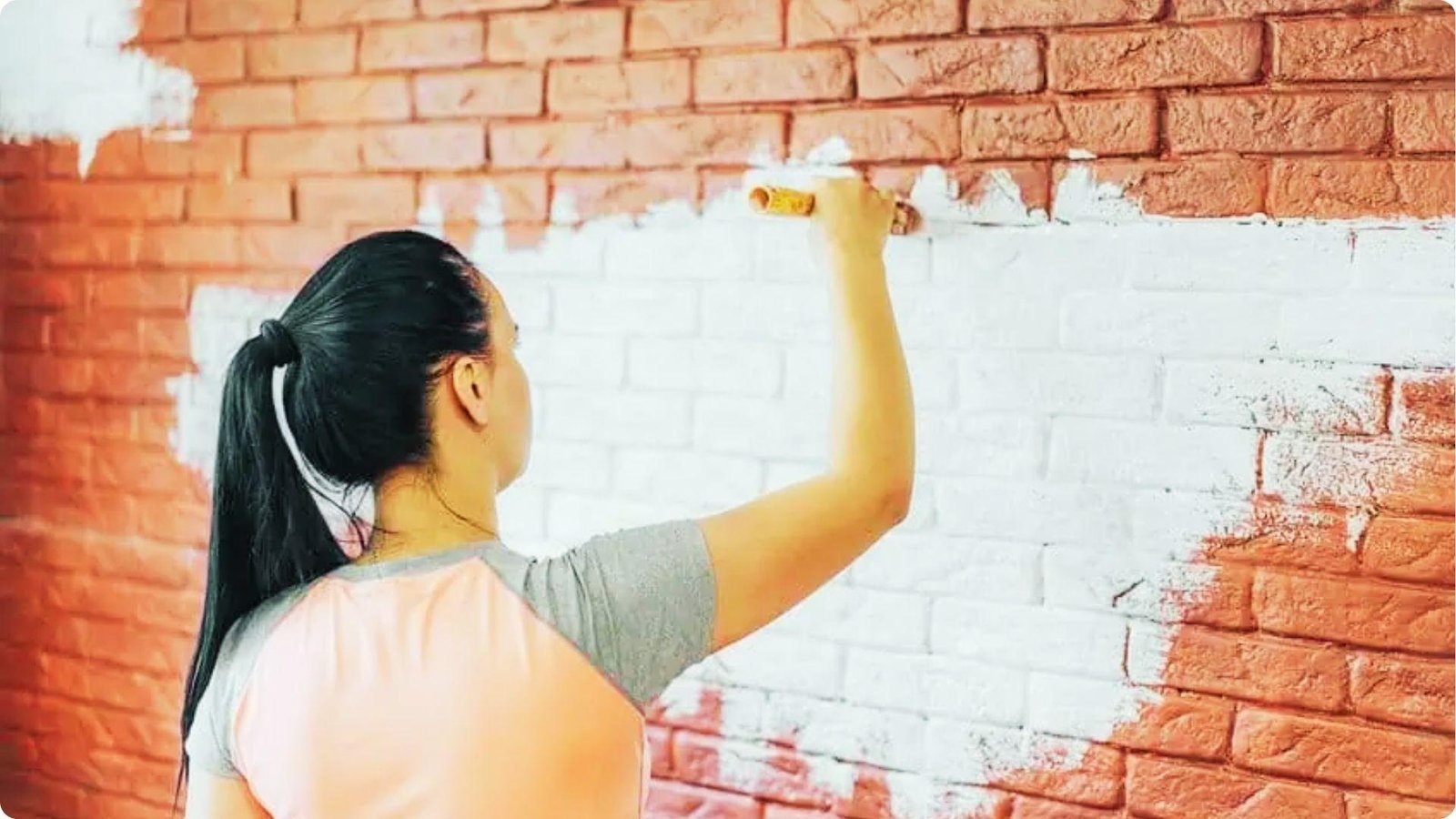 A woman with a ponytail uses a small roller to apply white paint onto a red brick wall.