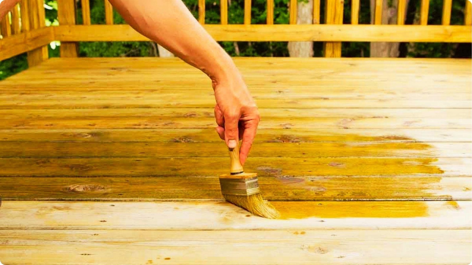 A hand uses a wide brush to apply golden stain to untreated deck boards.