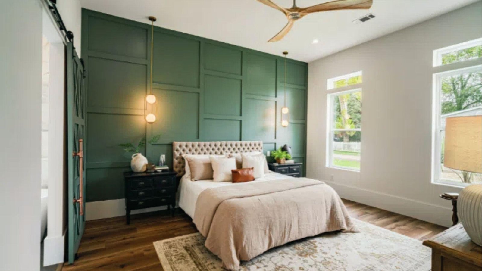 Modern bedroom with sage green paneled accent wall and light grey walls with barn door and fan.