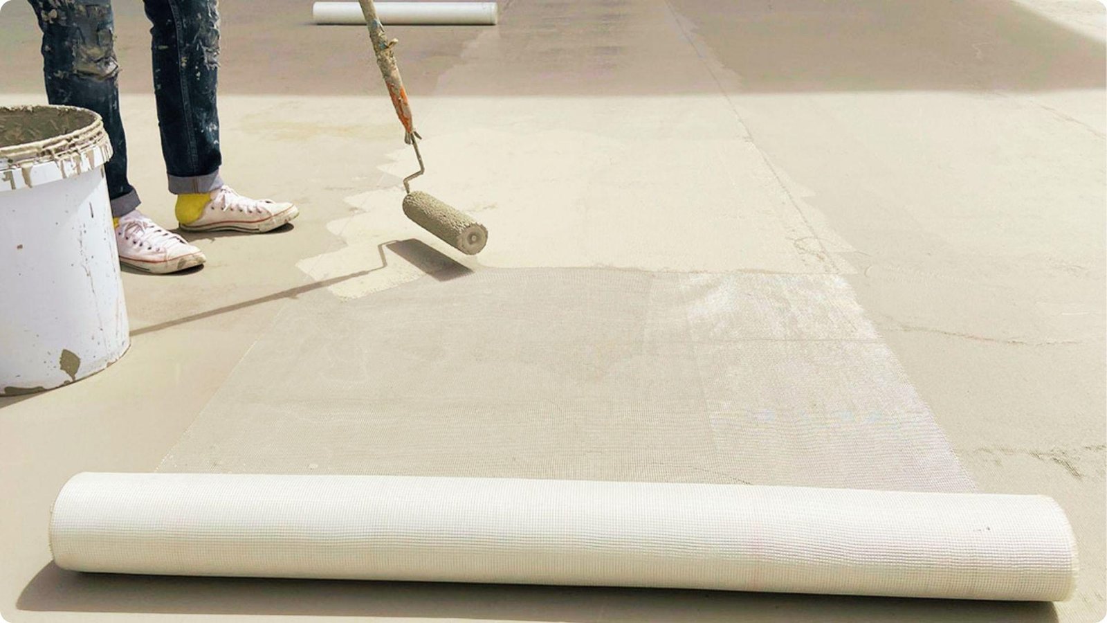 Worker in sneakers rolls beige coating over mesh fabric embedded into a flat roof, with a bucket nearby.