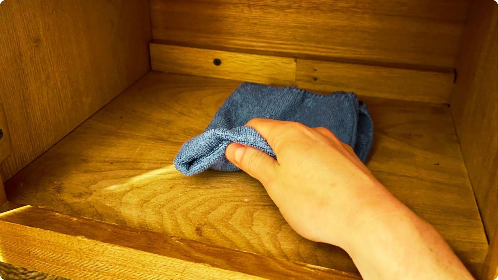 A hand wipes the inside of an empty wooden cabinet shelf with a blue microfiber cloth.