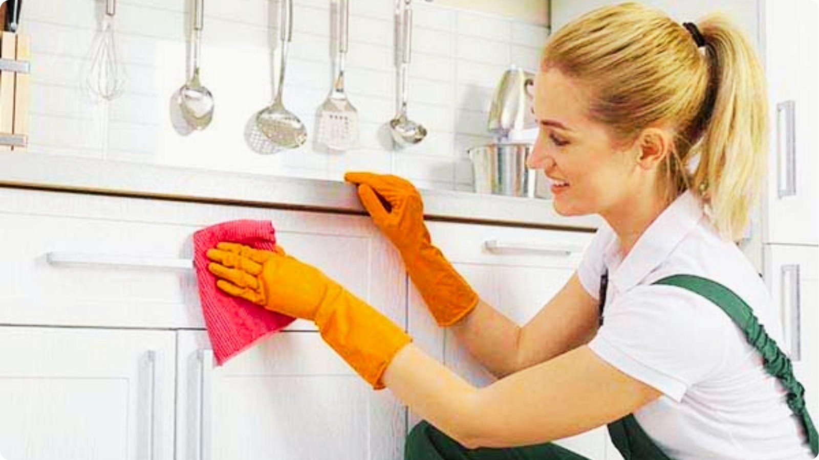 Woman wearing orange rubber gloves wipes down white painted kitchen cabinets with a pink microfiber cloth.