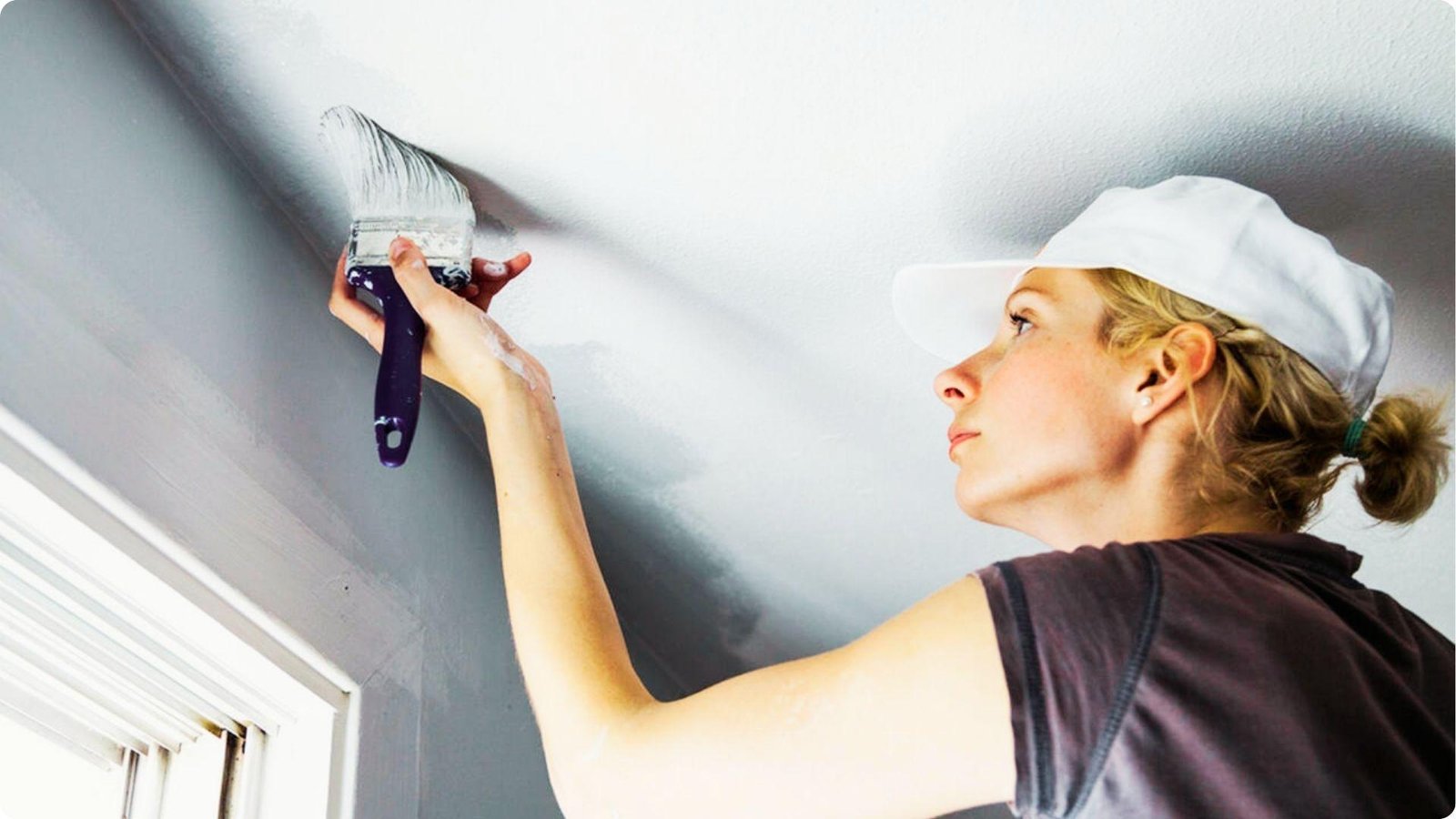Woman in a white cap uses a paintbrush to cut in white paint along the edge where a gray wall meets the ceiling.