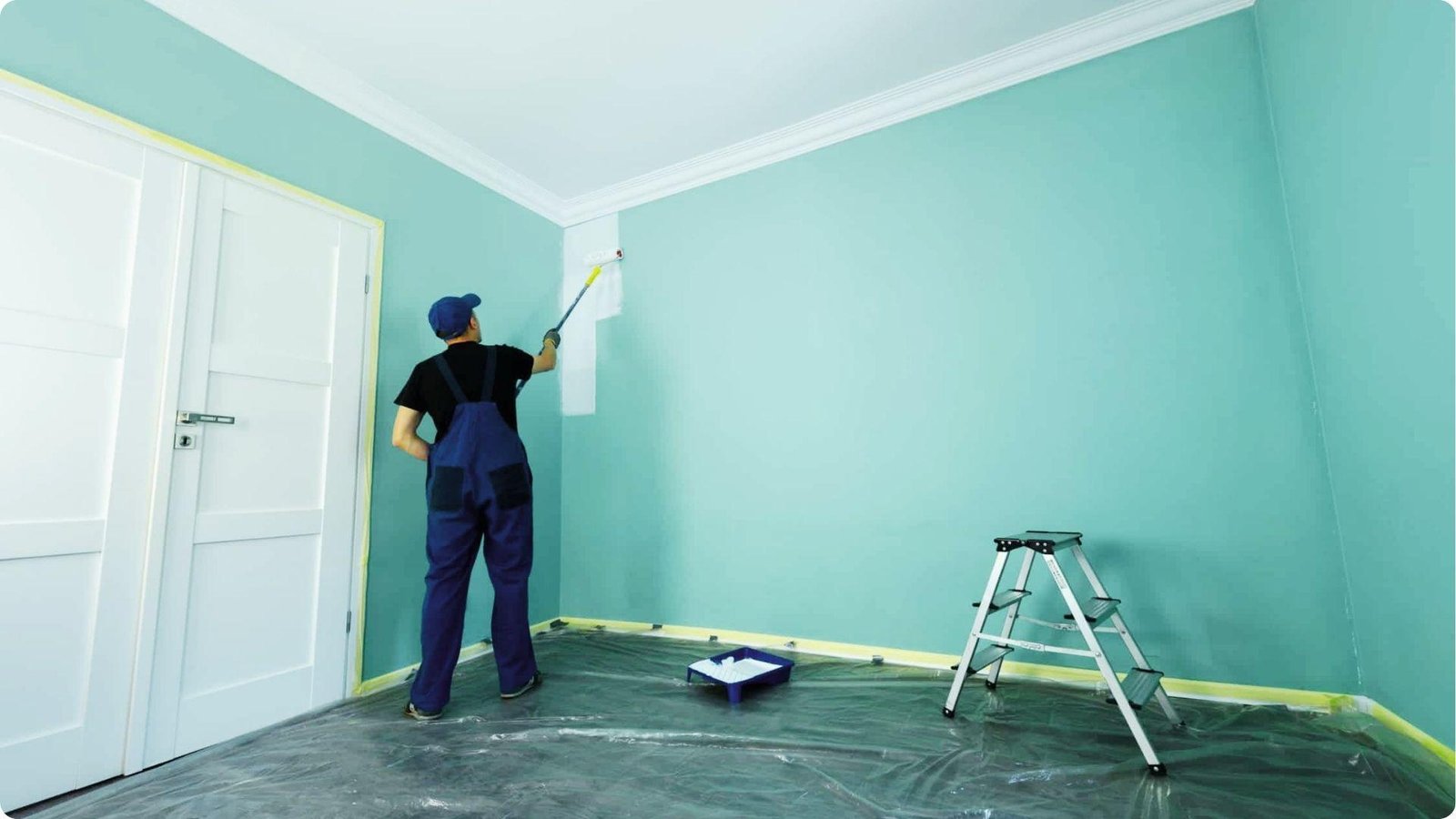 A professional in blue overalls and a cap rolls white paint over a teal wall in a room with floor protection and a step ladder.