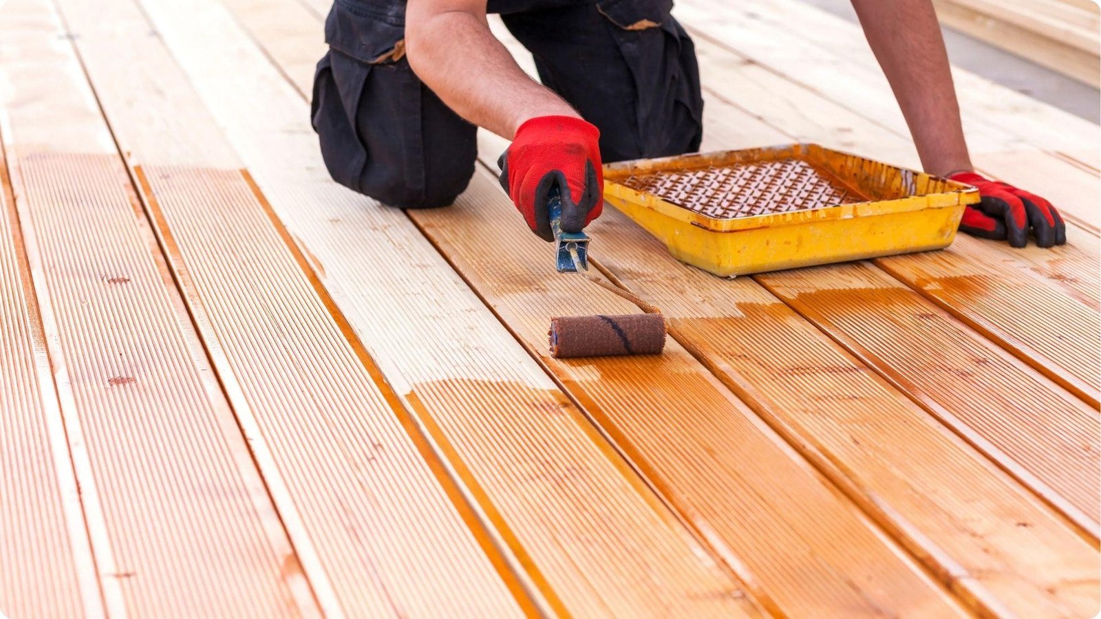 A person wearing red gloves rolls stain onto light-colored deck planks with a tray nearby.