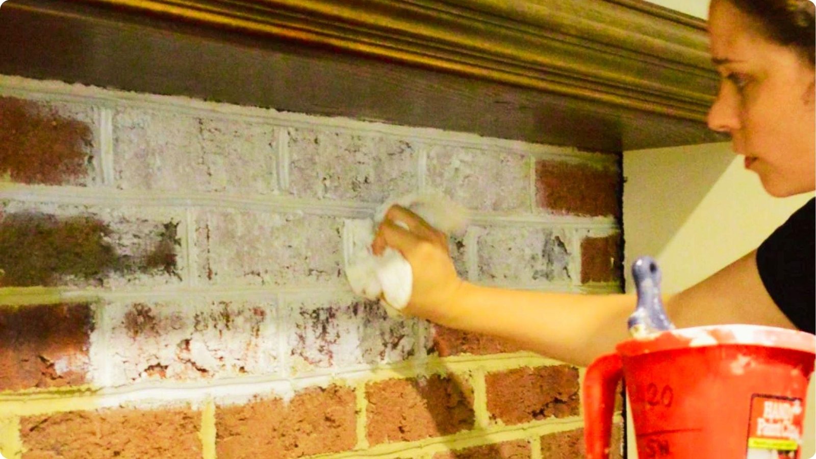 A woman uses a cloth to dab whitewash onto a red brick fireplace, creating a distressed look.