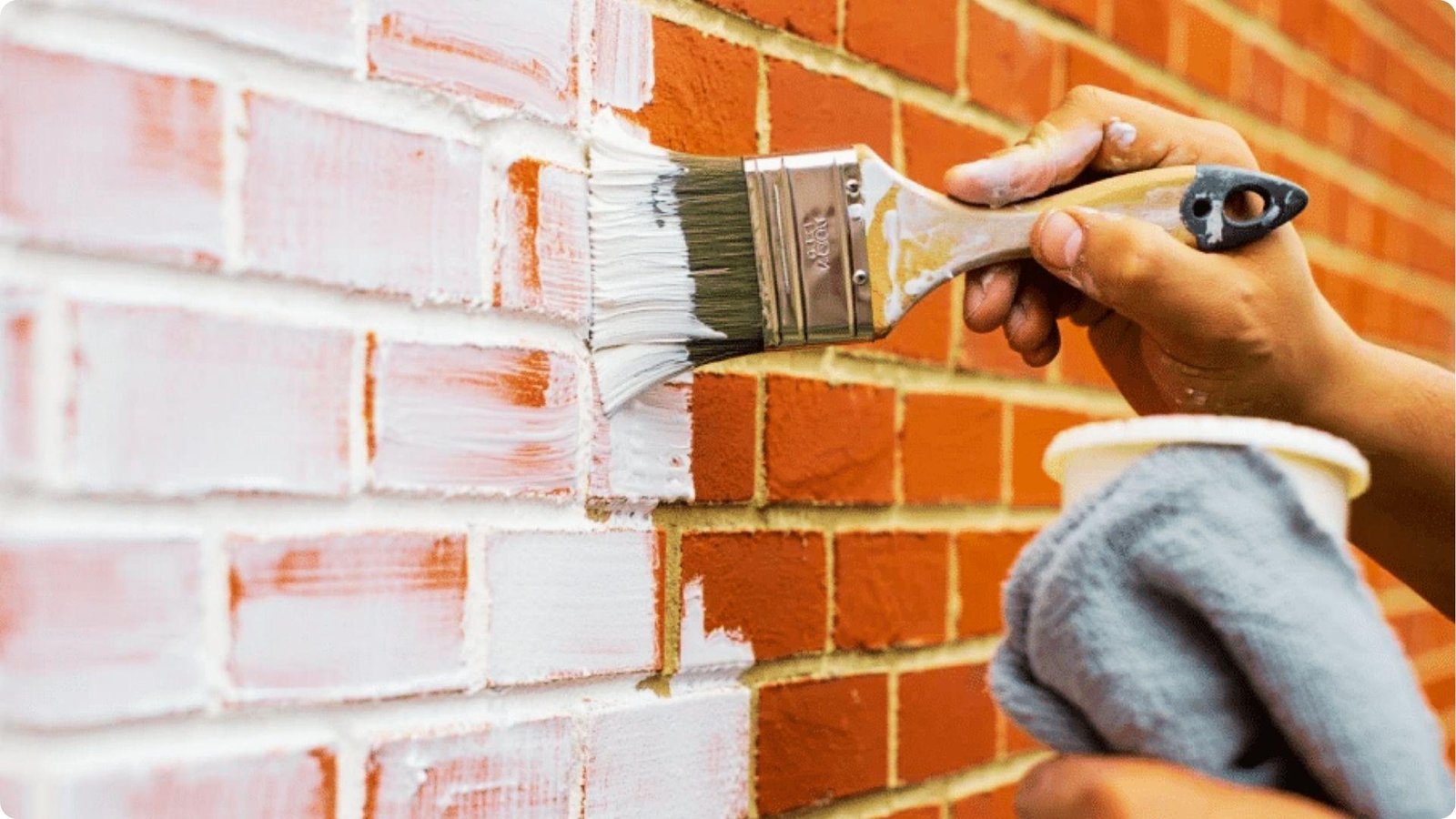 Close-up of a hand using a paintbrush to apply whitewash to red brick.