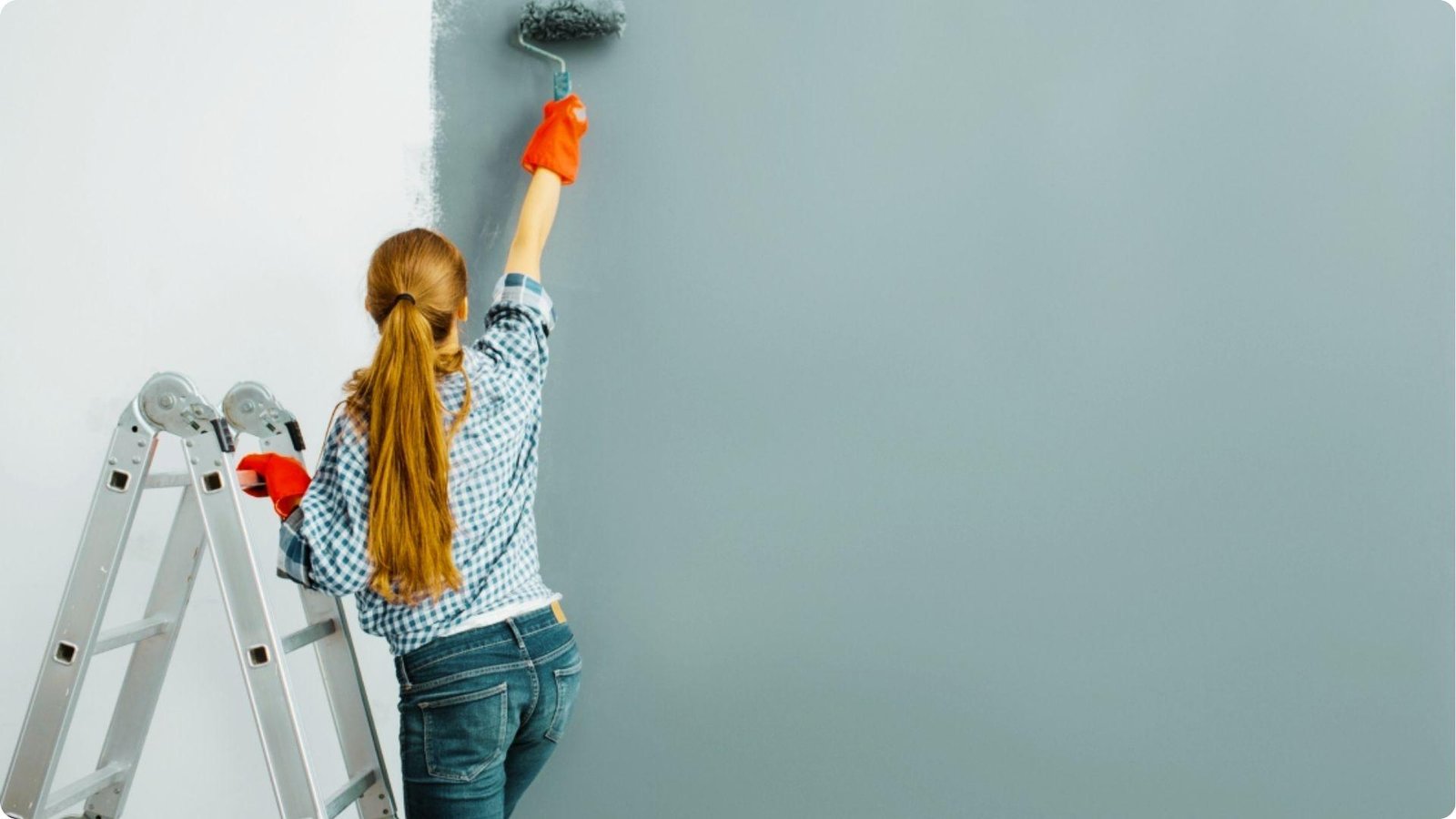 Woman in plaid shirt and orange gloves rolling gray paint onto a white wall from a ladder.