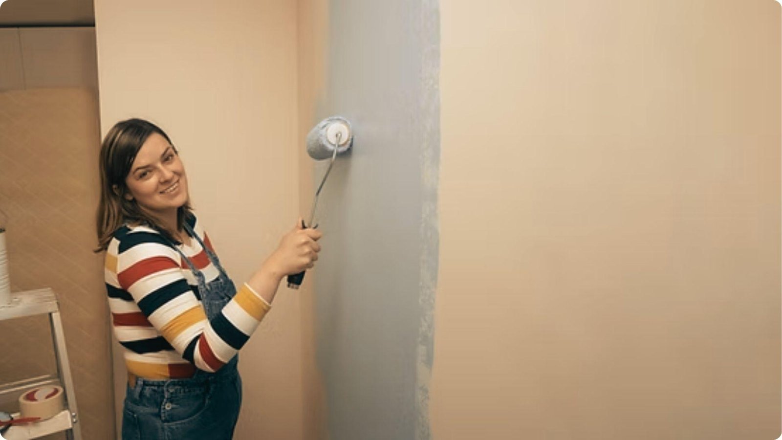 A woman in striped sleeves and denim overalls smiles as she rolls light gray paint over a beige wall.