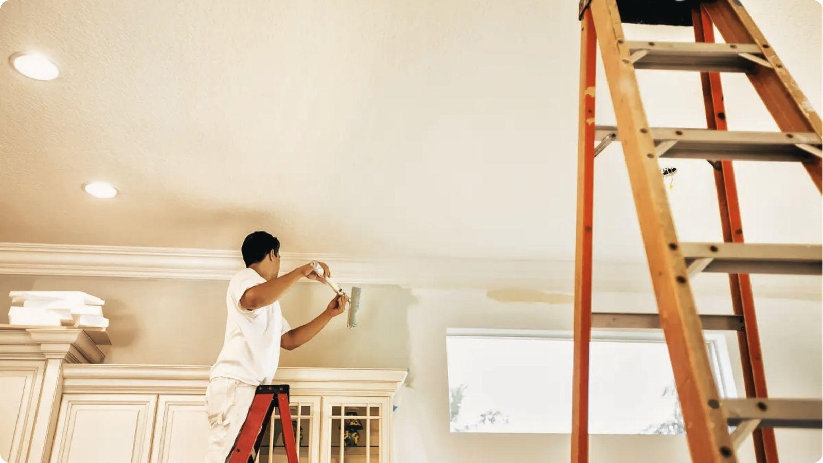 A professional painter in white attire uses a roller to paint an interior wall while standing on a red ladder.