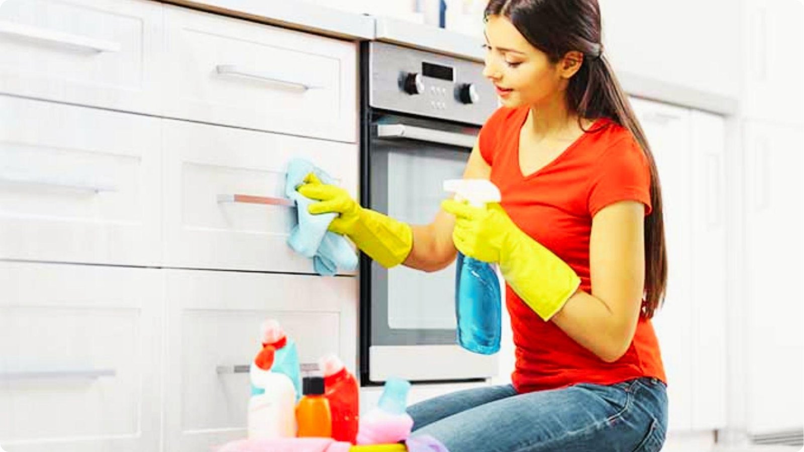 A woman in yellow rubber gloves sprays cleaning solution on a cloth to wipe down white painted kitchen drawers.