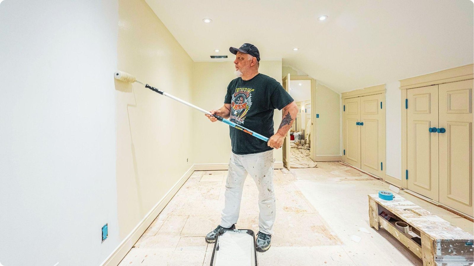 Professional painter in a cap using an extension roller to paint a cream-colored wall.