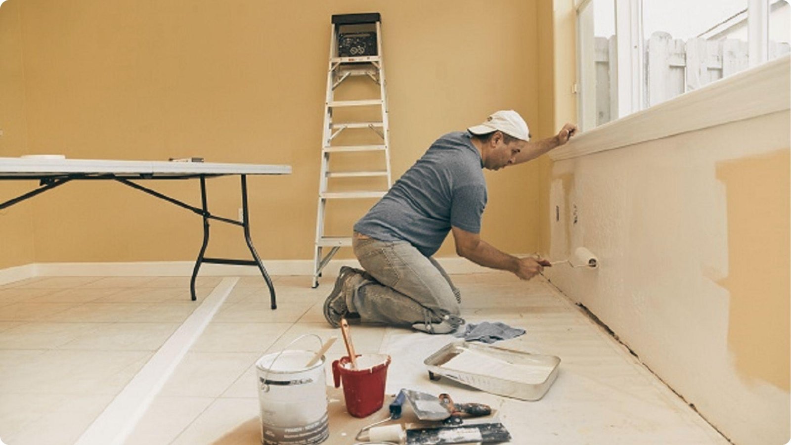 A painter in a cap kneels to apply paint near baseboards, with paint cans, trays, and a ladder around him.
