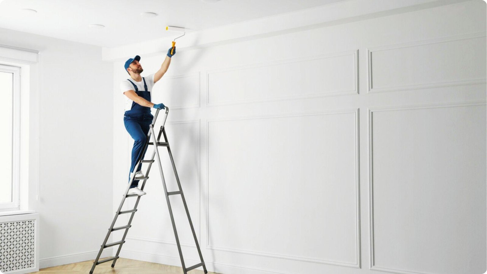 Man in blue overalls stands on a stepladder using a roller to paint a white ceiling near decorative wall paneling.