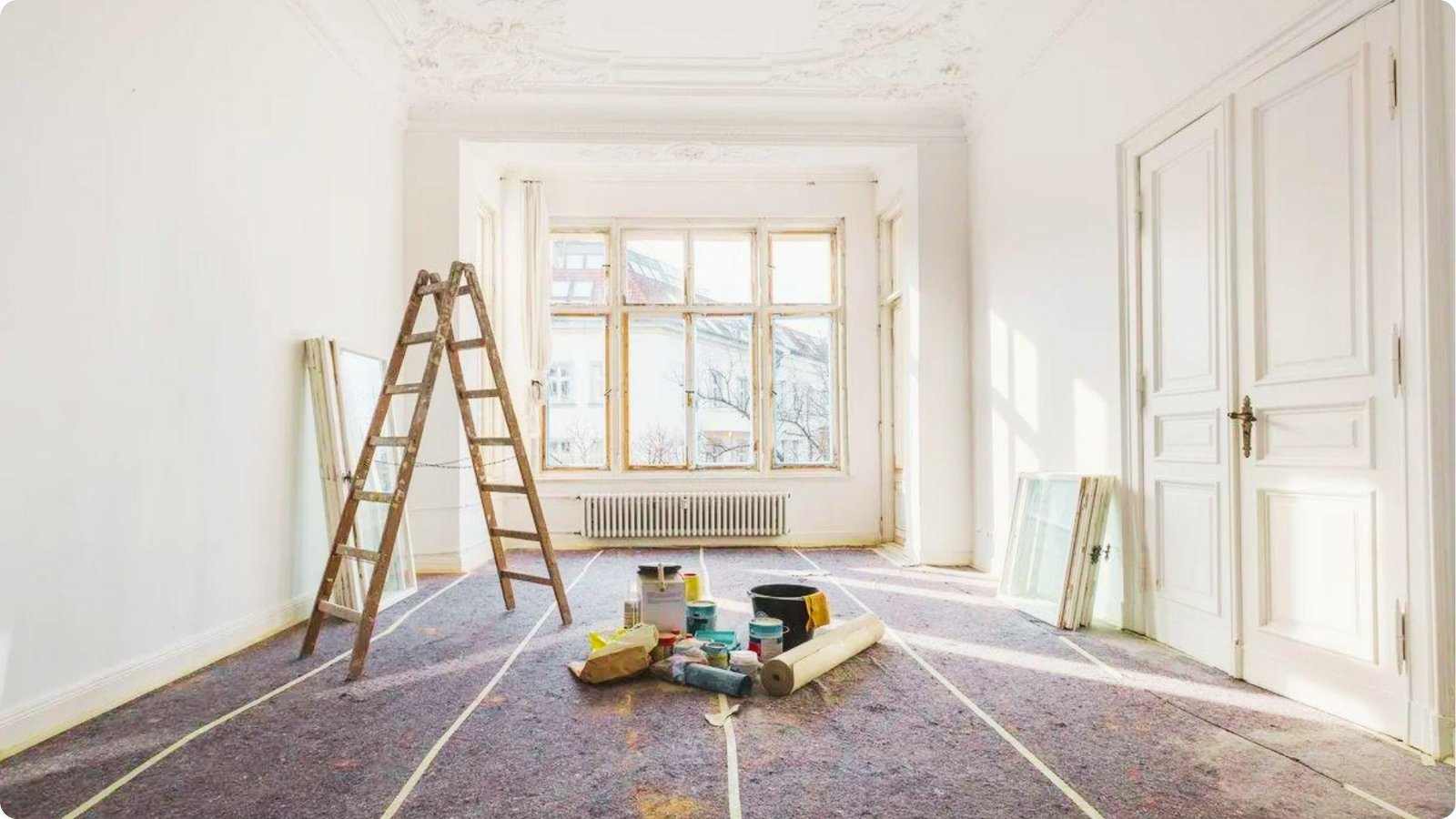Bright room prepped for painting with ladder, drop cloths, and paint cans near large windows.