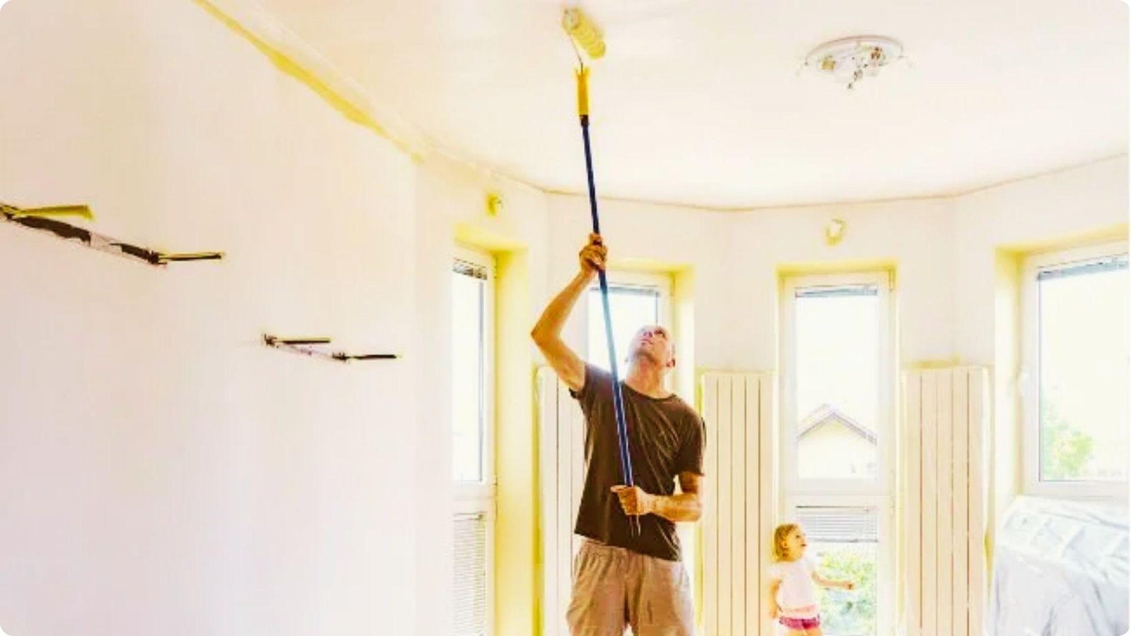 A man in a brown shirt uses an extension pole roller to apply yellow paint to a white ceiling while a child watches.
