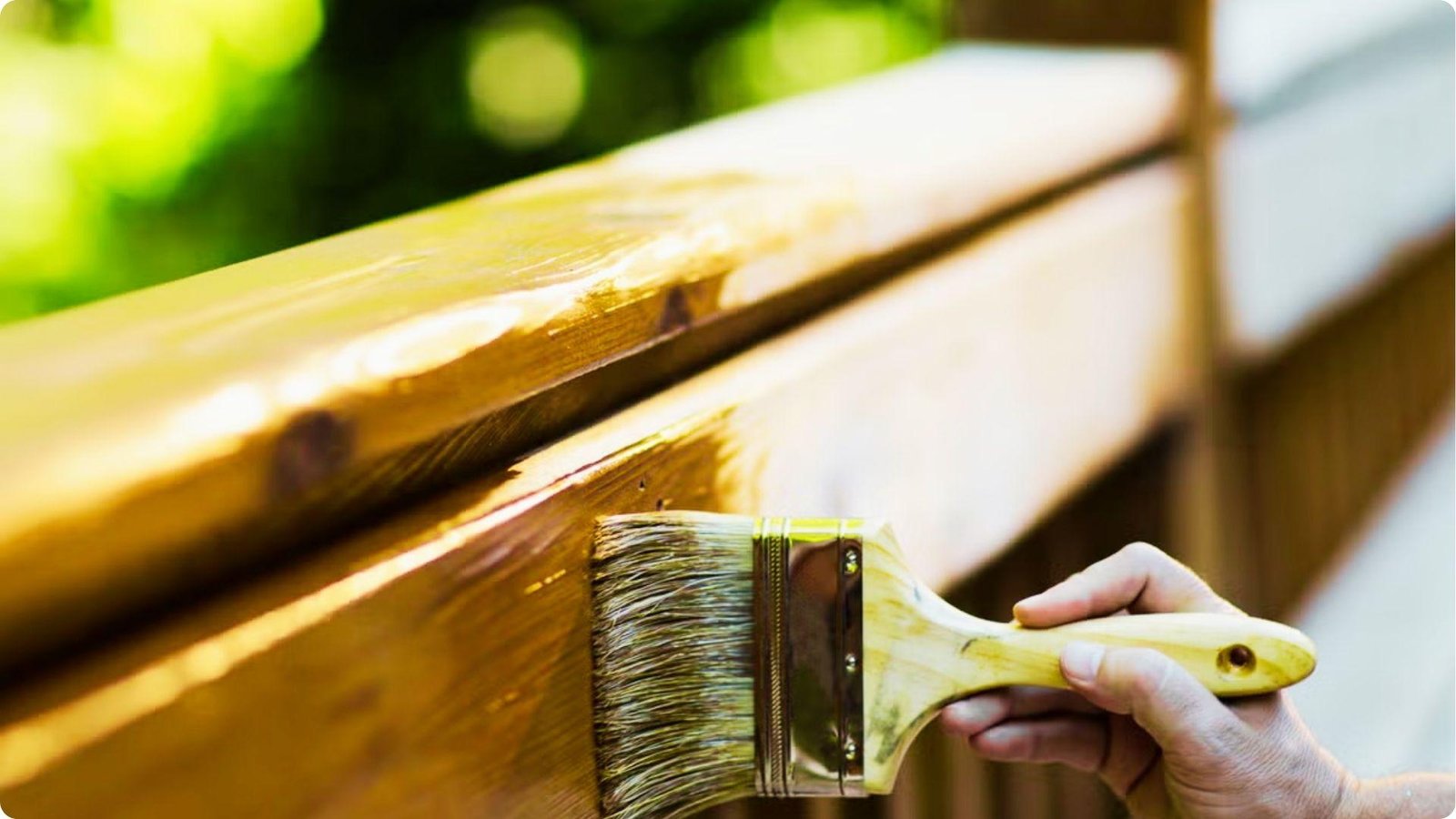 Close-up of a hand brushing amber-colored stain onto a deck railing with greenery behind it.