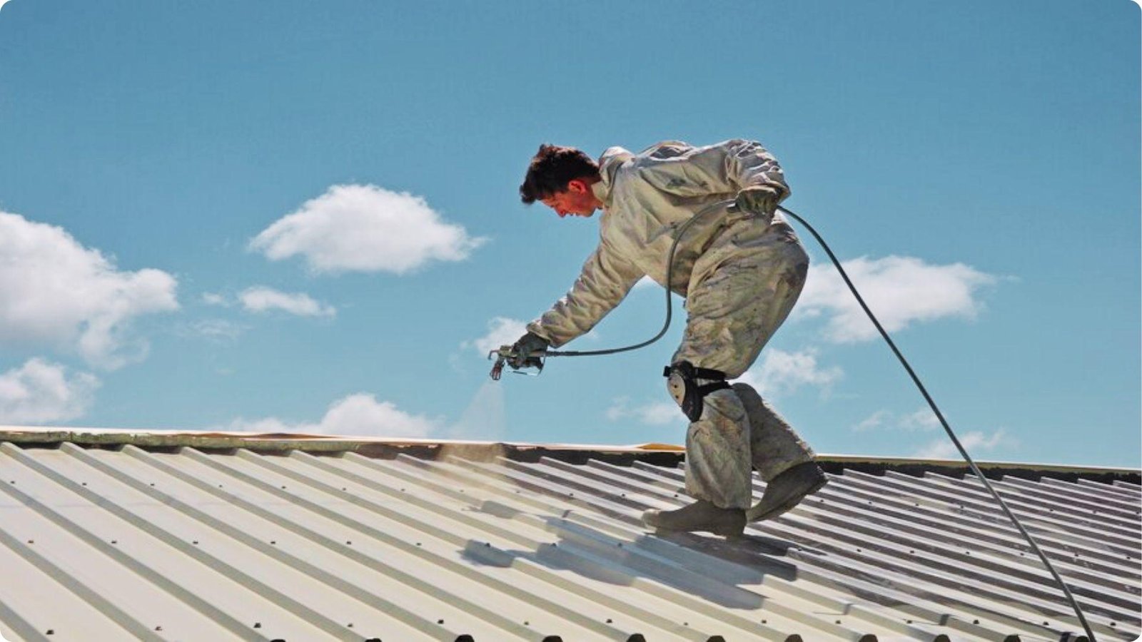 A worker in a white protective suit spray-coats a corrugated metal roof under a bright blue sky.