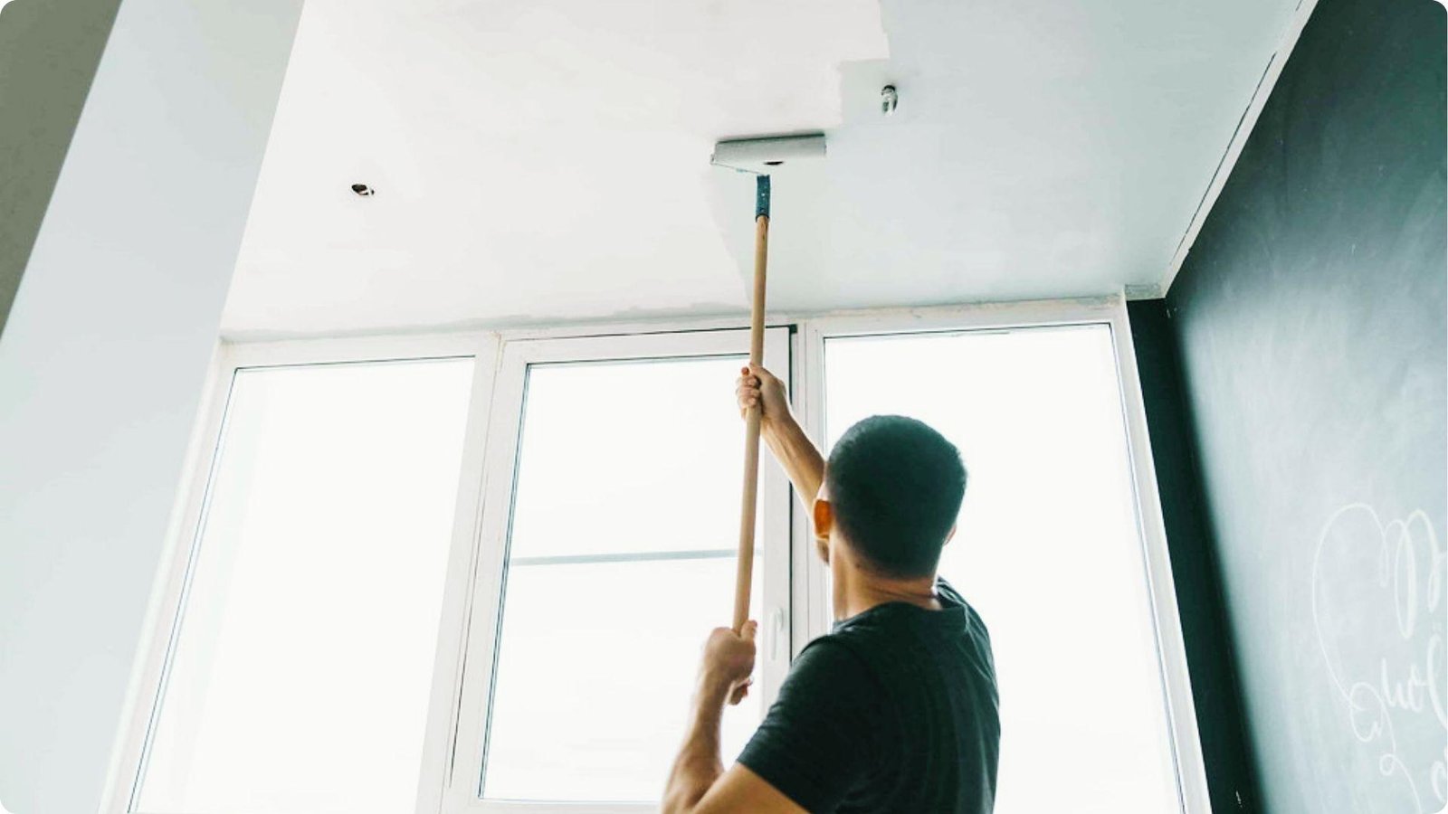 Person on a stepladder rolls white paint onto a ceiling next to a dark accent wall and large windows.