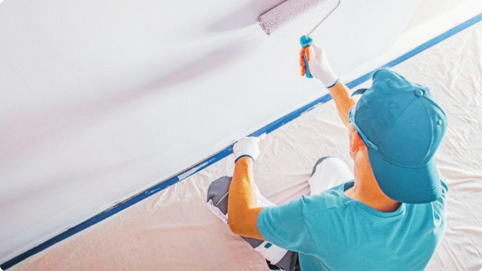 A painter wearing gloves and a cap uses a roller to paint a wall white, with blue painter's tape along the trim.