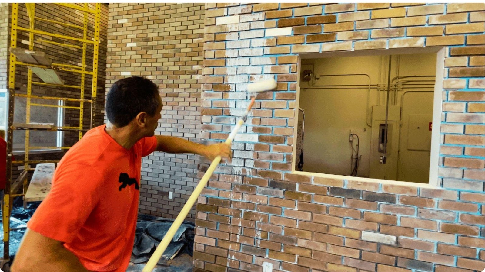 A man in an orange shirt uses a paint roller to apply whitewash to a large interior brick wall.