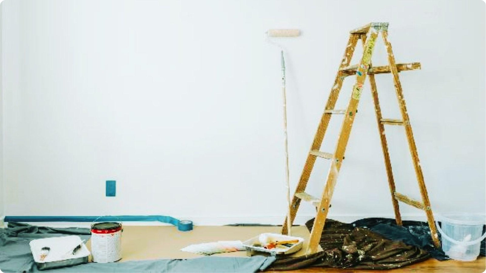 Painting setup with wooden ladder, roller, trays, and drop cloths against a blank white wall.