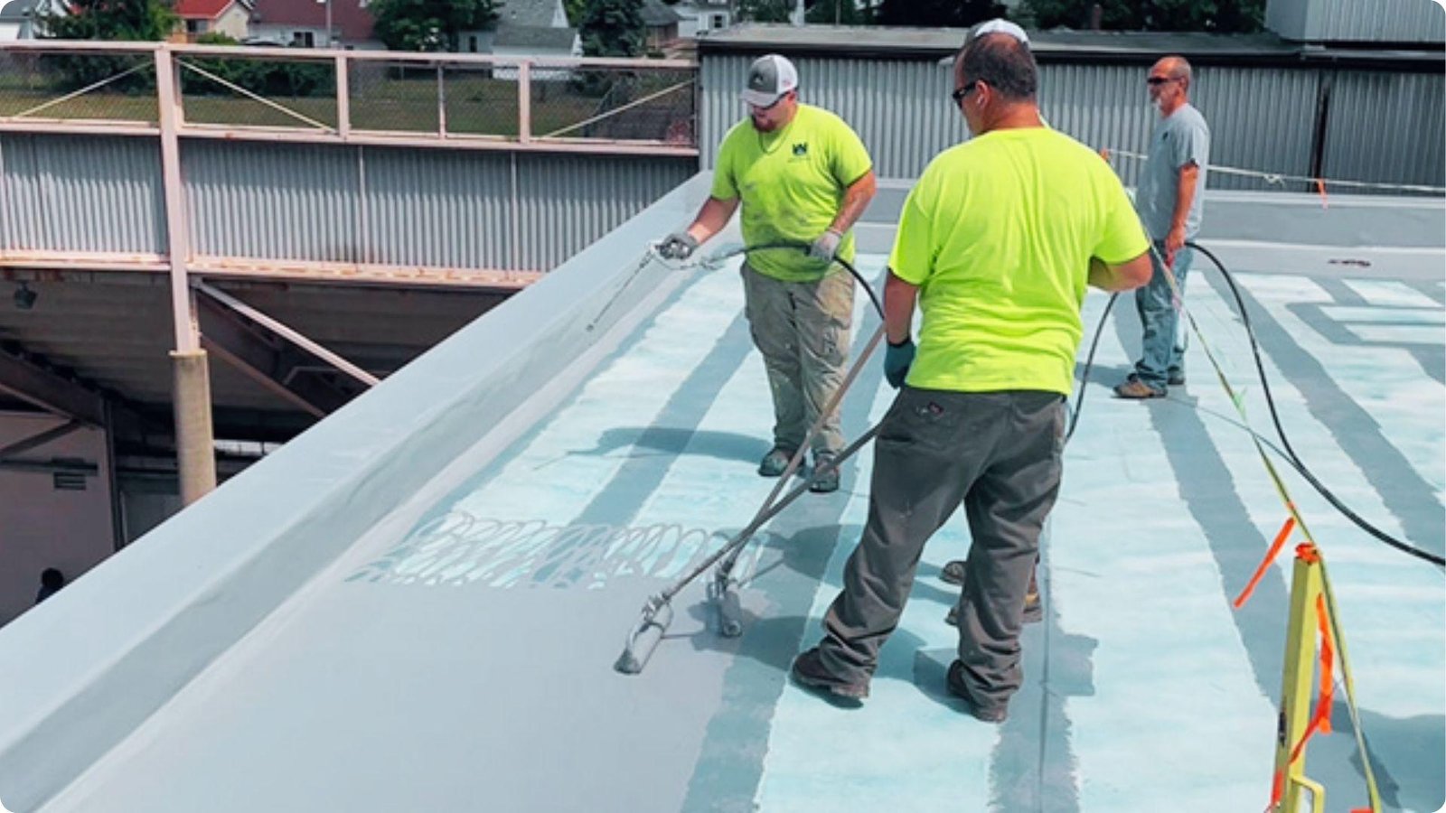 Three roofers in safety vests use spray equipment and rollers to apply a reflective coating across a large commercial TPO roof.