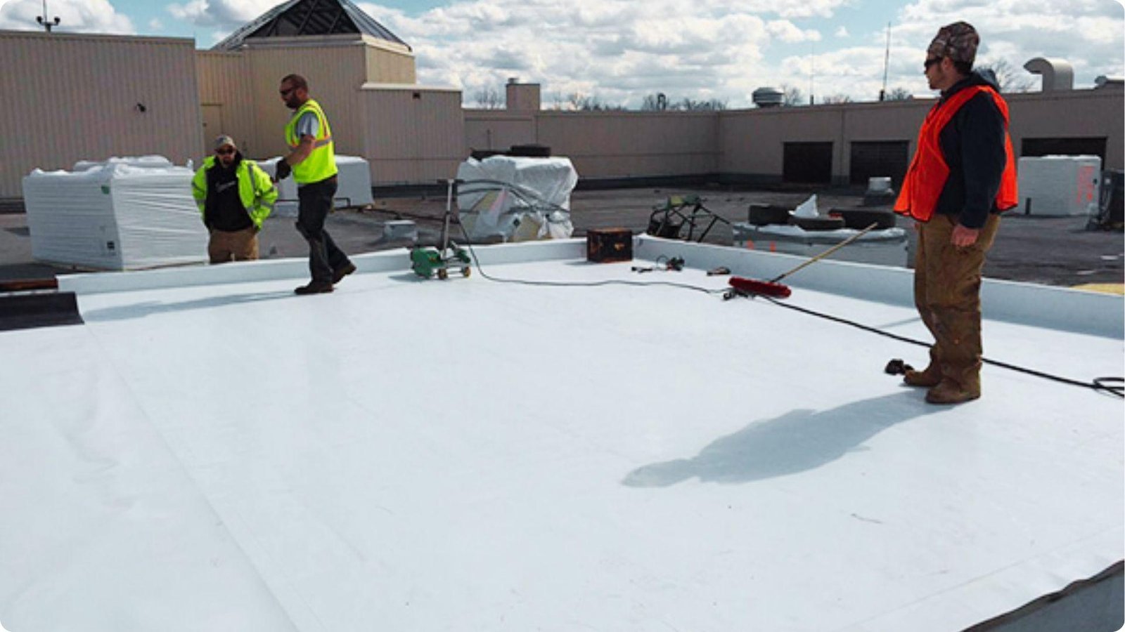 Crew in high-visibility vests installs and coats a bright white TPO roof membrane on a commercial building.