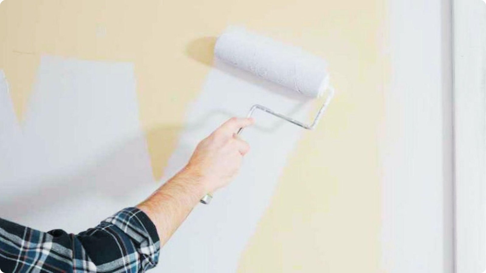 A hand in a plaid shirt uses a paint roller to apply a fresh coat of white paint over a pale yellow wall.