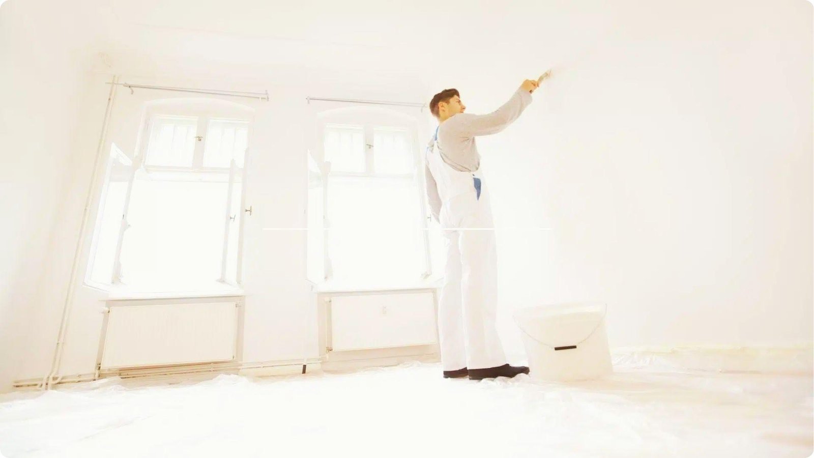 Painter in white overalls applies white paint to an interior wall with a brush in a bright, empty room.